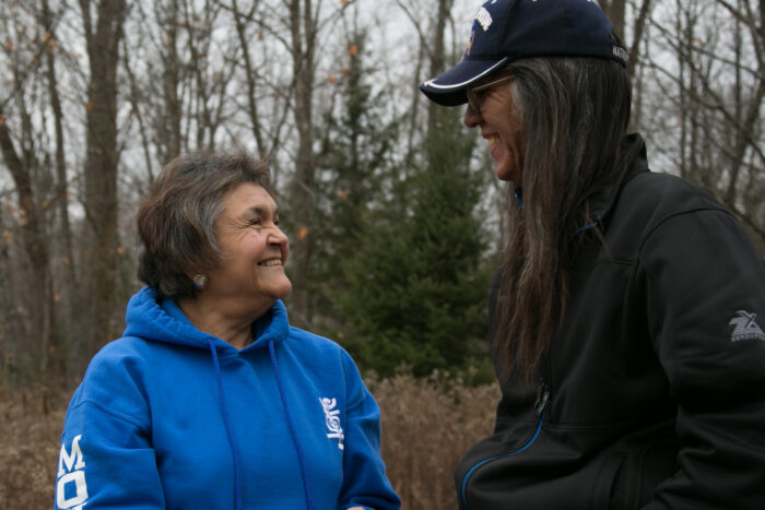 A color photograph showing Molly Miller and another person standing in front of a wooded area. They are looking at each other and are smiling. Molly wears a blue hooded shirt with the word 