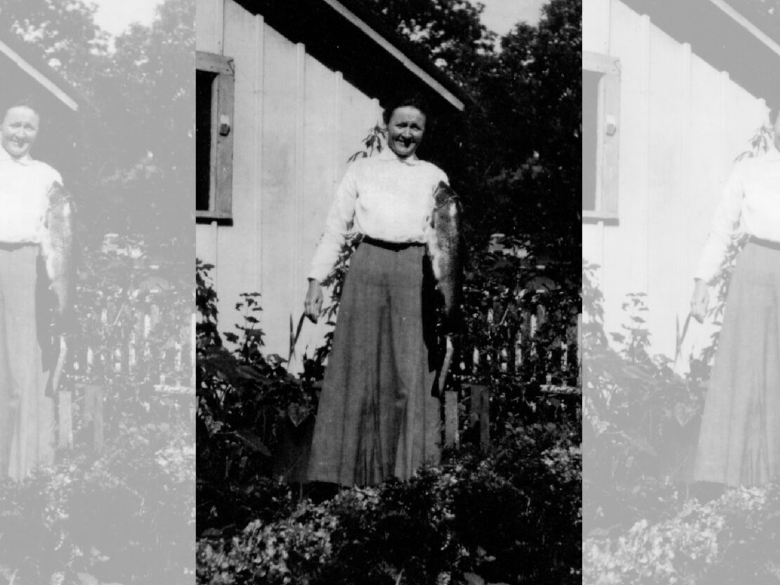 A black and white photo of Carrie standing in front of a building holding up a fish.