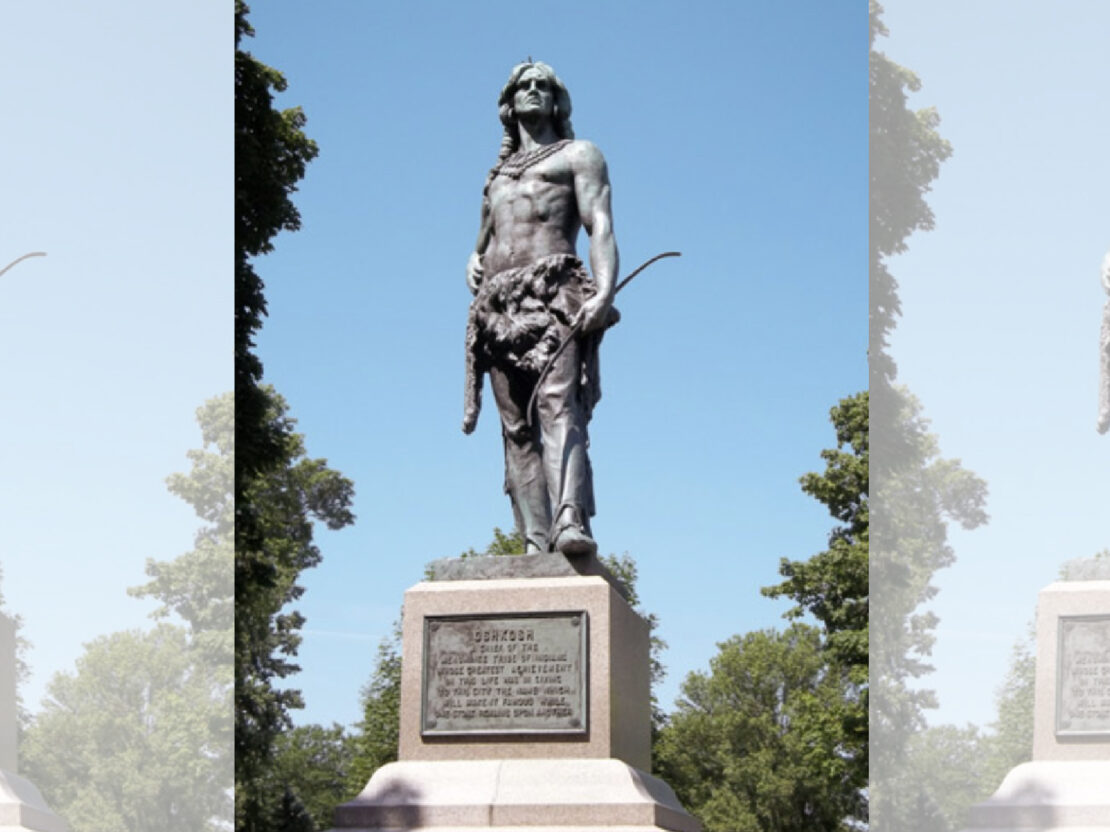 A statue of a man standing with his shoulders back and arms near his sides, on top of a square cement base that has a metal plaque on it.