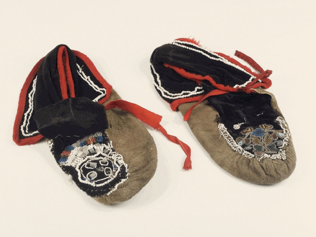 A color photograph of a pair of moccasins. They are tan and black with red accents and multicolored beadwork.