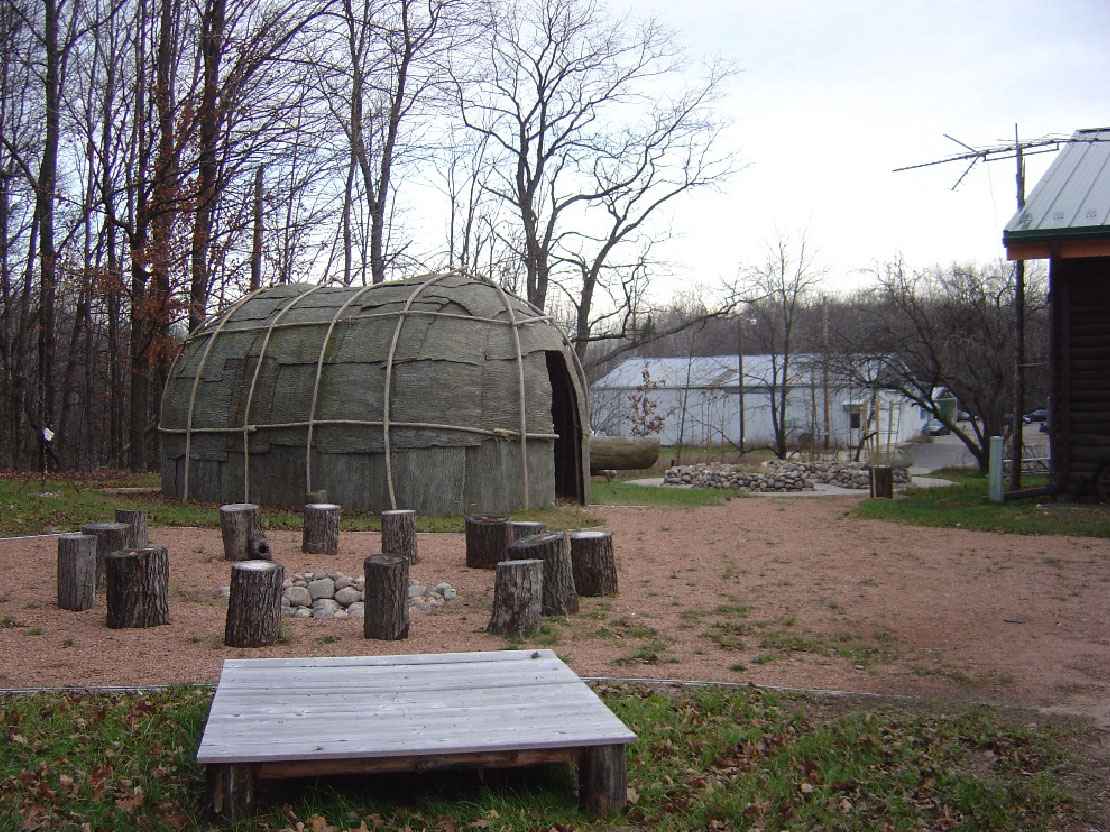 A photograph of a modern recreation of a wigwam. It is an oval structure originally made of wooden poles and tree bark.