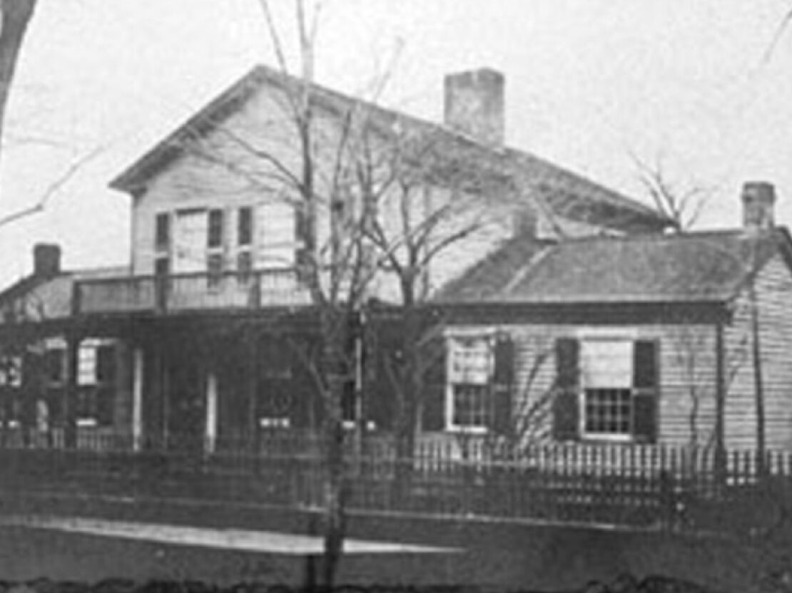 A black and white photograph of a white house with a porch, framed by trees.