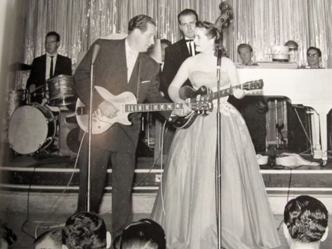 A black and white photograph of Les Paul and Mary Ford performing on stage, each holding a guitar, with a large band behind them.