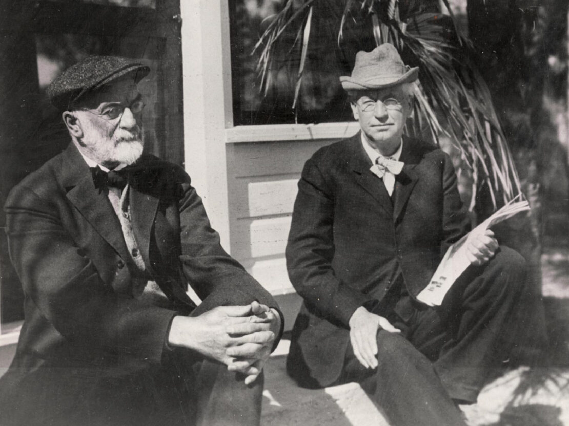 A black and white image showing two older men seated on a porch.