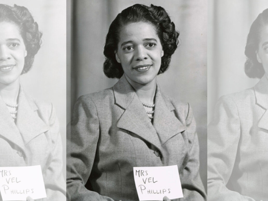 A black and white photograph of Vel Phillips. In the photo, Vel is looking to the right of the viewer and holds a piece of paper with the words “Mrs. Vel Phillips” on it.