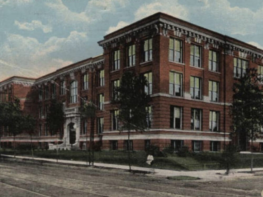 A color illustration of North Division High School. The illustration is from street level, and shows a large red brick building with light gray accents, against a light blue sky with some clouds.