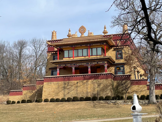 A color photograph of a large Buddhist temple. The temple is made of sand-colored stones and accented with maroon, bright red, and gold. There are large gold ornamental sculptures on the roof.