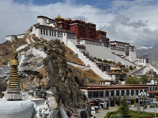 A photograph of a large set of buildings on a mountain. The lower walls are white and the upper walls are maroon. In the distance are more mountains.