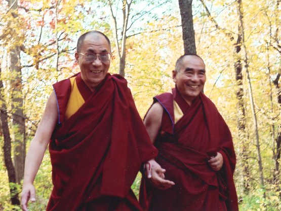 A color photograph of Geshe Sopa and the Dalai Lama walking together. They are both wearing maroon monastic robes. Behind them are trees with yellow and green leaves.