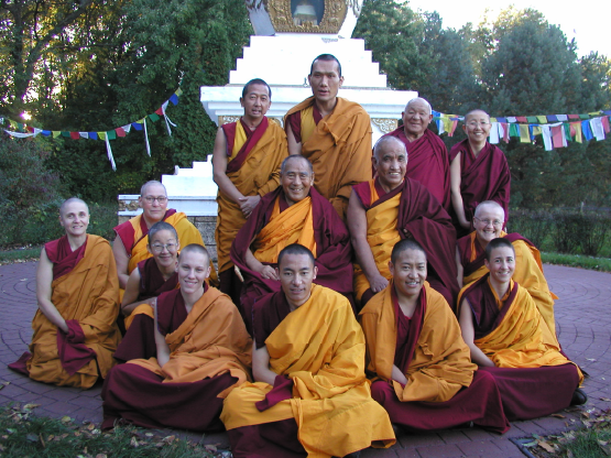 A color photograph of a group of monks and nuns. They are seated and standing outside in front of a white and gold shrine. The monks all wear maroon and yellow robes. Geshe Sopa is seated in the center of the group.