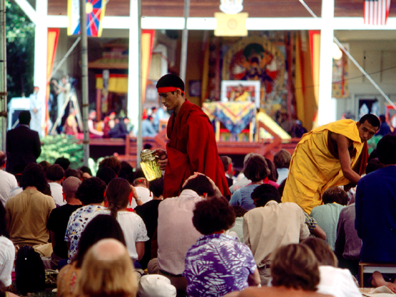 A color photograph of a group of people seated and looking toward a large, brightly decorated pavilion. Near the center of the image a young monk in maroon robes walks by holding a pitcher of tea.