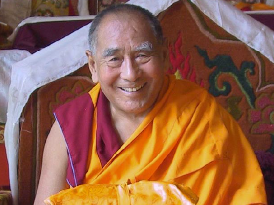 A color photograph of Geshe Lhundub Sopa as an older man. He is seated and smiling. He wears his monastic robes. In front of him is an object wrapped in yellow fabric.
