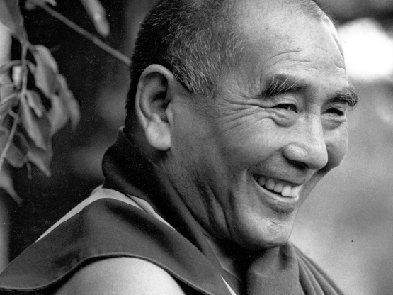 A black and white photograph of Geshe Lhundub Sopa. In the photograph, his head and shoulders are visible and he is smiling while looking towards the viewer's right.