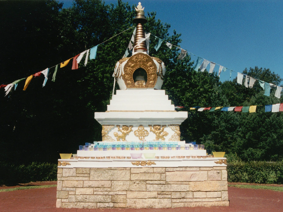 A color photograph of a Buddhist stupa: there is a sand-colored square stone base, on top of which is a white and gold structure that gets narrow towards the top. Strands of multicolored prayer flags are attached to the top of the structure.
