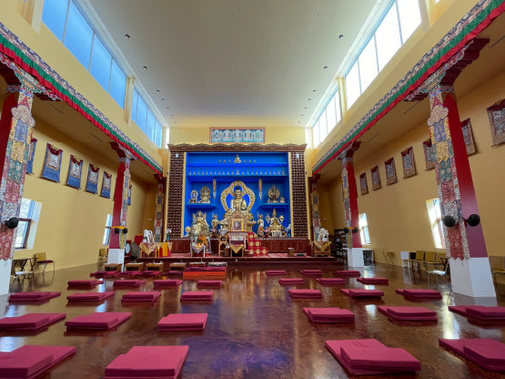 A color photograph of the interior of a Buddhist temple. The walls are yellow with decorative tapestries hanging up high. At the far end is a bright blue altar wall with a large gold statue of the Buddha and other gold decorations. Maroon mats are arranged in rows for seating on the floor.
