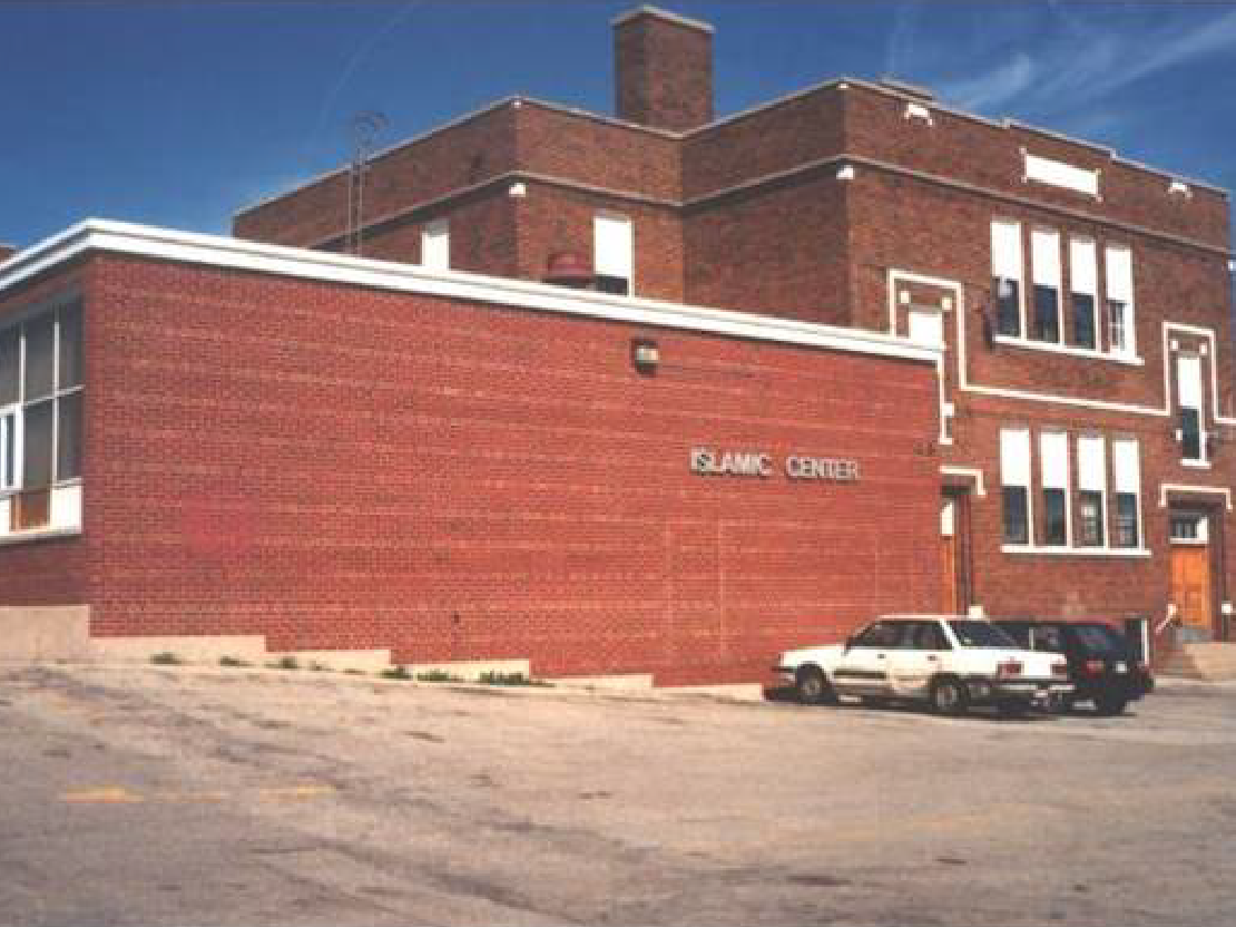 A photograph of an old school building. It is a brick building with some large windows. One wall is entirely brick, without windows, and has a sign that says "Islamic Center."